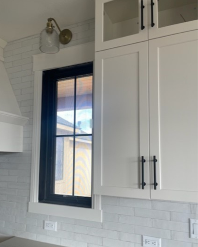 Bright kitchen wall with white shaker cabinets and black hardware, a small window with dark trim, white subway tile backsplash, and a wall-mounted lamp with a brass shade above the window.